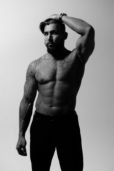 Hispanic shirtless male model with muscular tattooed torso standing with hands in pockets and looking away on gray backdrop