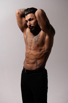 Hispanic shirtless male model with muscular tattooed torso standing with hands in pockets and looking away on gray backdrop