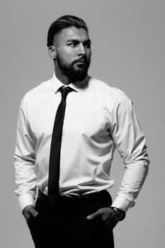 Bearded Hispanic guy in dark vest and white shirt with tie looking at camera with hands in pockets in studio