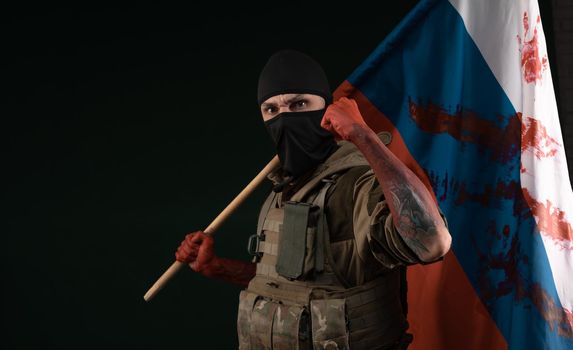 male soldier in a military uniform with a balaclava and hands covered in blood holds a bloody flag of Russia on a black background