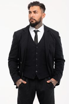 Bearded Hispanic guy in dark vest and white shirt with tie looking at camera with hands in pockets in studio