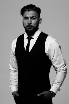 Bearded Hispanic guy in dark vest and white shirt with tie looking at camera with hands in pockets in studio