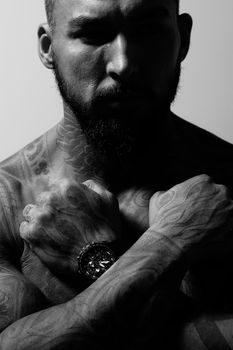 Hispanic shirtless male model with muscular tattooed torso standing with hands in pockets and looking away on gray backdrop