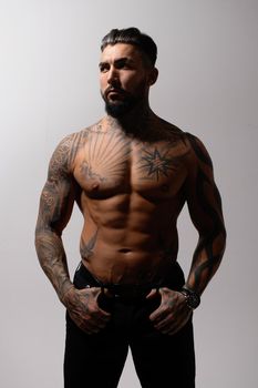 Hispanic shirtless male model with muscular tattooed torso standing with hands in pockets and looking away on gray backdrop