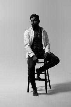 Hispanic shirtless male model with muscular tattooed torso standing with hands in pockets and looking away on gray backdrop