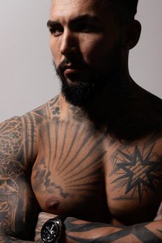 Hispanic shirtless male model with muscular tattooed torso standing with hands in pockets and looking away on gray backdrop