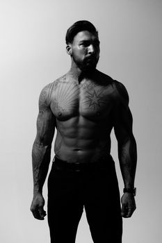 Hispanic shirtless male model with muscular tattooed torso standing with hands in pockets and looking away on gray backdrop