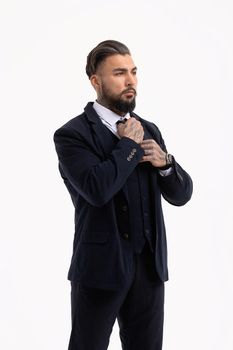 Confident Hispanic bearded man with tattooed hands in white shirt and black trousers adjusting tie and looking away while standing against gray background