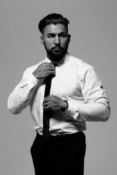 Confident Hispanic bearded man with tattooed hands in white shirt and black trousers adjusting tie and looking away while standing against gray background