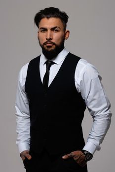 Bearded Hispanic guy in dark vest and white shirt with tie looking at camera with hands in pockets in studio