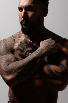 Hispanic shirtless male model with muscular tattooed torso standing with hands in pockets and looking away on gray backdrop