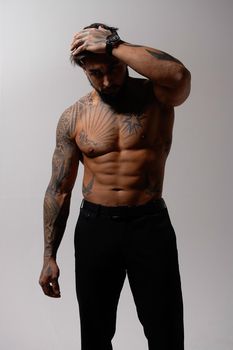 Hispanic shirtless male model with muscular tattooed torso standing with hands in pockets and looking away on gray backdrop