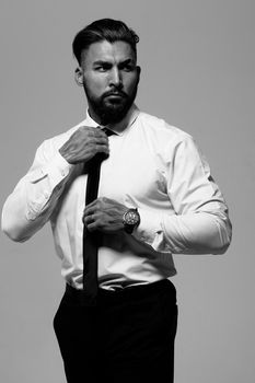 Confident Hispanic bearded man with tattooed hands in white shirt and black trousers adjusting tie and looking away while standing against gray background