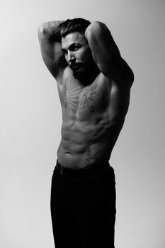 Hispanic shirtless male model with muscular tattooed torso standing with hands in pockets and looking away on gray backdrop