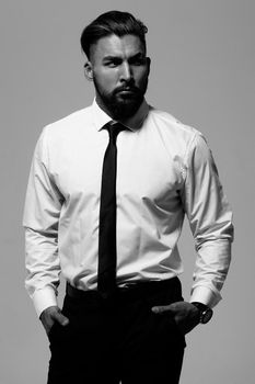 Bearded Hispanic guy in dark vest and white shirt with tie looking at camera with hands in pockets in studio