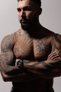 Hispanic shirtless male model with muscular tattooed torso standing with hands in pockets and looking away on gray backdrop