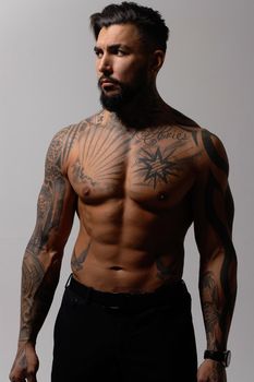 Hispanic shirtless male model with muscular tattooed torso standing with hands in pockets and looking away on gray backdrop