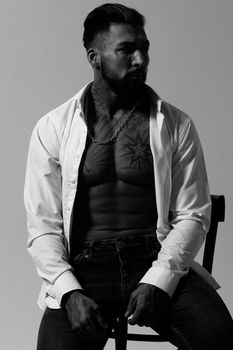 Hispanic shirtless male model with muscular tattooed torso standing with hands in pockets and looking away on gray backdrop