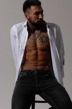 Hispanic shirtless male model with muscular tattooed torso standing with hands in pockets and looking away on gray backdrop