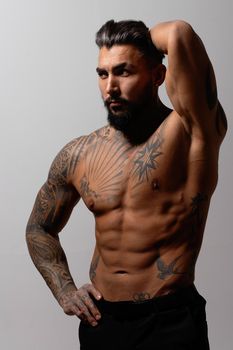 Hispanic shirtless male model with muscular tattooed torso standing with hands in pockets and looking away on gray backdrop