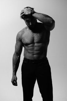 Hispanic shirtless male model with muscular tattooed torso standing with hands in pockets and looking away on gray backdrop