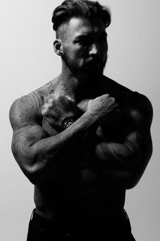 Hispanic shirtless male model with muscular tattooed torso standing with hands in pockets and looking away on gray backdrop