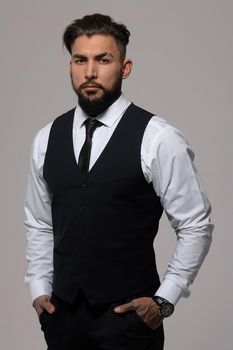 Bearded Hispanic guy in dark vest and white shirt with tie looking at camera with hands in pockets in studio