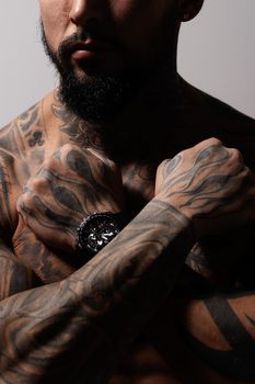 Hispanic shirtless male model with muscular tattooed torso standing with hands in pockets and looking away on gray backdrop