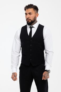 Bearded Hispanic guy in dark vest and white shirt with tie looking at camera with hands in pockets in studio