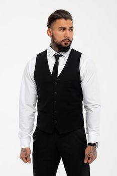 Bearded Hispanic guy in dark vest and white shirt with tie looking at camera with hands in pockets in studio