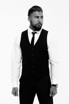 Bearded Hispanic guy in dark vest and white shirt with tie looking at camera with hands in pockets in studio