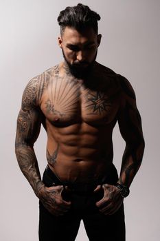 Hispanic shirtless male model with muscular tattooed torso standing with hands in pockets and looking away on gray backdrop