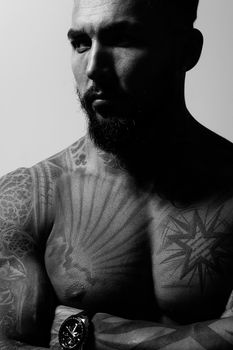 Hispanic shirtless male model with muscular tattooed torso standing with hands in pockets and looking away on gray backdrop