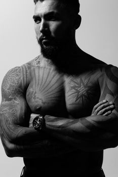 Hispanic shirtless male model with muscular tattooed torso standing with hands in pockets and looking away on gray backdrop