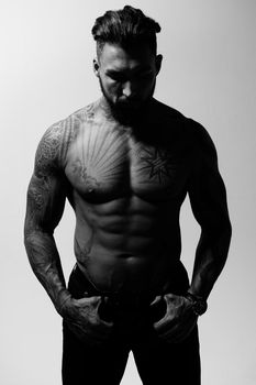 Hispanic shirtless male model with muscular tattooed torso standing with hands in pockets and looking away on gray backdrop