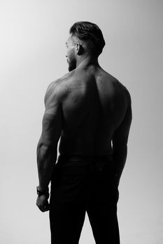 Hispanic shirtless male model with muscular tattooed torso standing with hands in pockets and looking away on gray backdrop