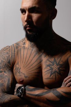 Hispanic shirtless male model with muscular tattooed torso standing with hands in pockets and looking away on gray backdrop