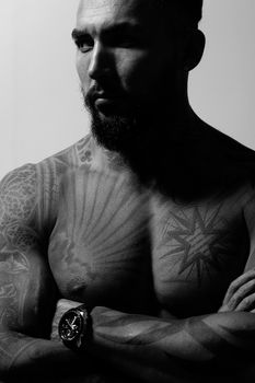 Hispanic shirtless male model with muscular tattooed torso standing with hands in pockets and looking away on gray backdrop