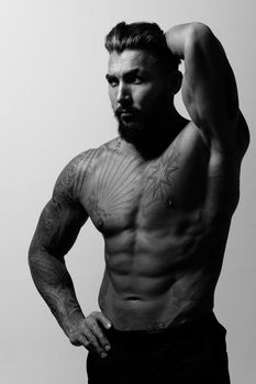 Hispanic shirtless male model with muscular tattooed torso standing with hands in pockets and looking away on gray backdrop