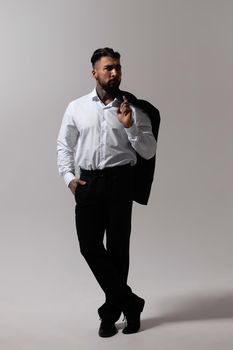 Bearded Hispanic guy in dark vest and white shirt with tie looking at camera with hands in pockets in studio