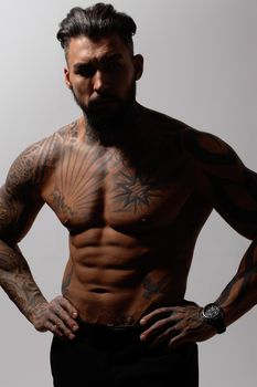 Hispanic shirtless male model with muscular tattooed torso standing with hands in pockets and looking away on gray backdrop