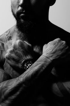 Hispanic shirtless male model with muscular tattooed torso standing with hands in pockets and looking away on gray backdrop