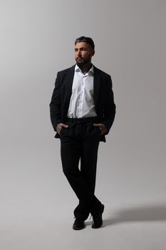 Bearded Hispanic guy in dark vest and white shirt with tie looking at camera with hands in pockets in studio