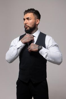 Confident Hispanic bearded man with tattooed hands in white shirt and black trousers adjusting tie and looking away while standing against gray background