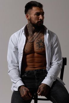 Hispanic shirtless male model with muscular tattooed torso standing with hands in pockets and looking away on gray backdrop