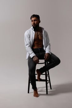 Hispanic shirtless male model with muscular tattooed torso standing with hands in pockets and looking away on gray backdrop