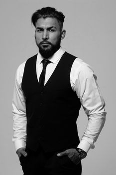 Bearded Hispanic guy in dark vest and white shirt with tie looking at camera with hands in pockets in studio