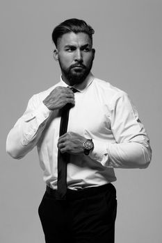 Confident Hispanic bearded man with tattooed hands in white shirt and black trousers adjusting tie and looking away while standing against gray background