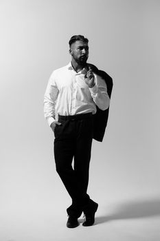 Bearded Hispanic guy in dark vest and white shirt with tie looking at camera with hands in pockets in studio