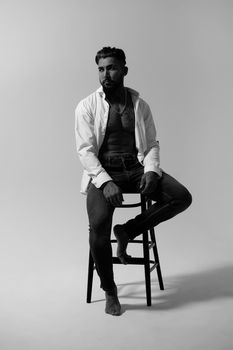 Hispanic shirtless male model with muscular tattooed torso standing with hands in pockets and looking away on gray backdrop